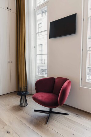 Una habitación con una silla roja, un televisor en la pared y una ventana con vistas a edificios.