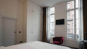 A hotel room with a bed, a red chair, a TV, and large windows. - Riga Suites | Deluxe Double Room with Bath (Antwerpen)