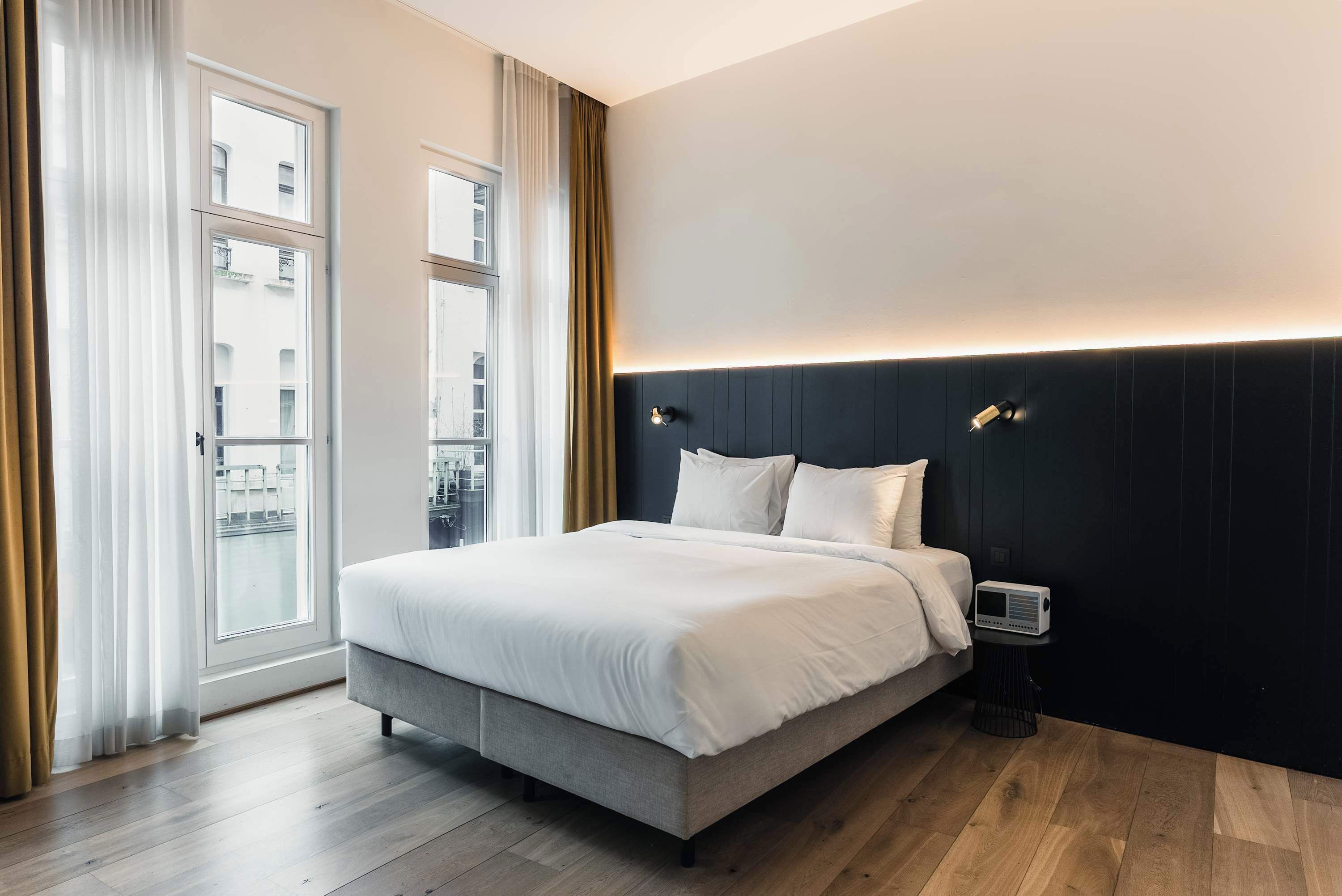 Ein modernes Hotelzimmer mit einem großen Bett, einem Nachttisch, einem Fenster mit Vorhängen und Blick auf die Stadt.