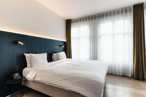 Una habitación de hotel moderna con una cama grande, un teléfono en la mesita de noche y una ventana con cortinas ligeras.