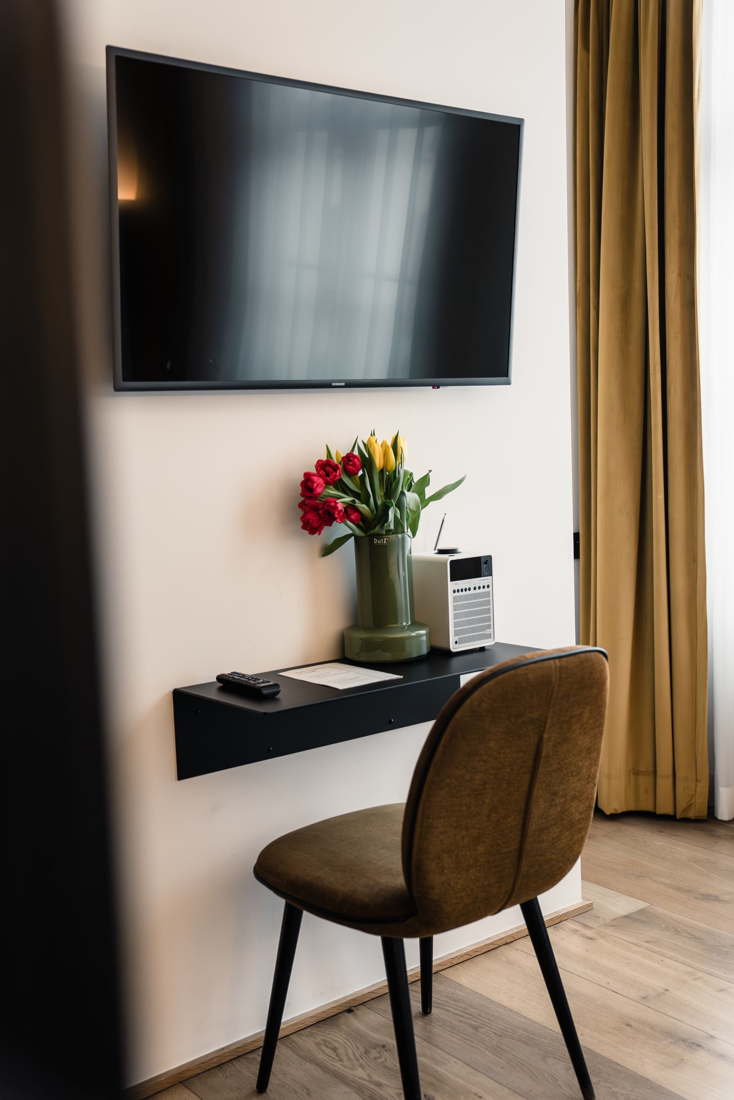 Una habitación moderna con televisor de pantalla plana montado en la pared, una mesita con un control remoto, un florero con flores y una silla.