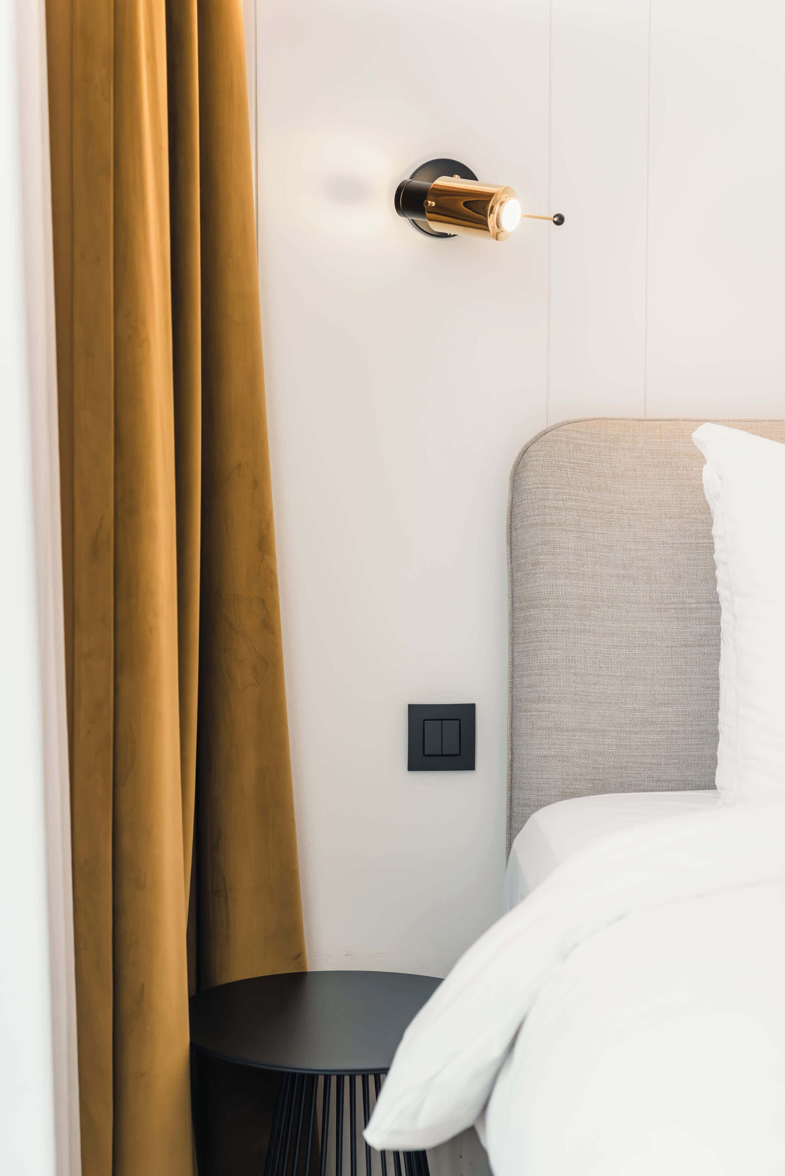 A hotel room with a bed, a bedside table, a wall-mounted light fixture, and a curtain.