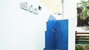 Eine blaue Duschkabine mit sichtbarer Brause, eine weiße Wand mit einer angebrachten Leuchte und ein Holzzaun mit Grünpflanzen.