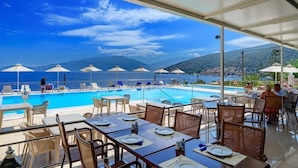 A poolside dining area with tables and chairs, umbrellas, and a view of the sea and mountains. - Odyssey Hotel (Kefalonia)