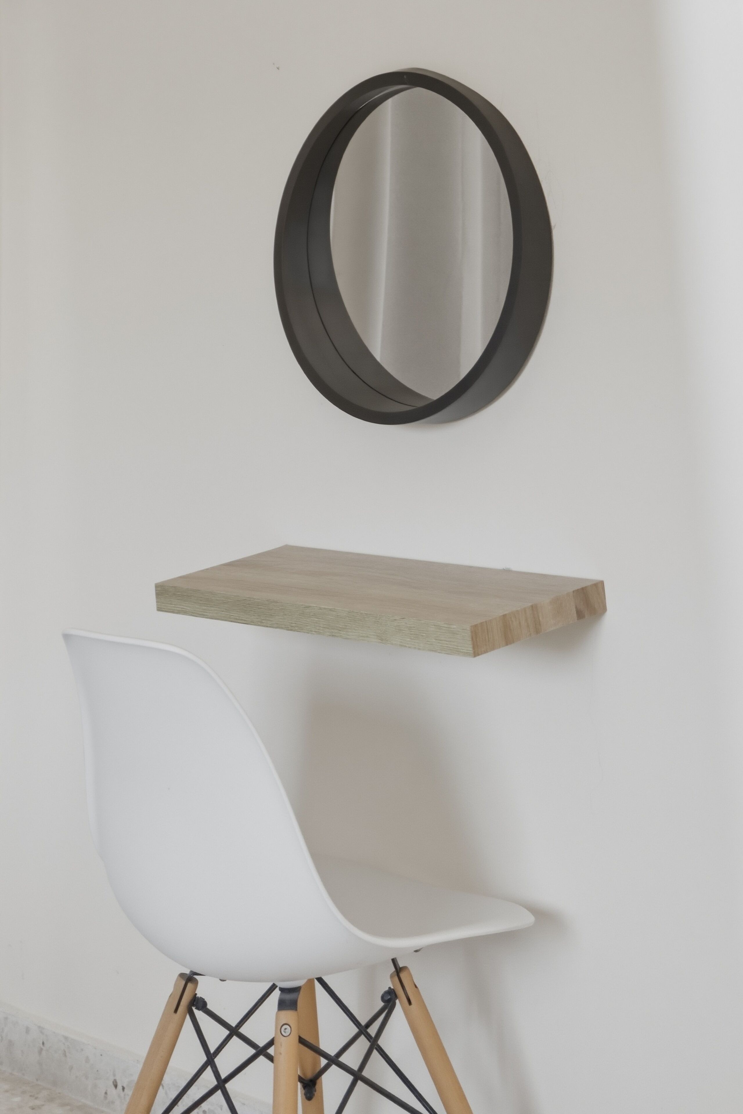 A white chair with a wooden base, a round mirror on a wall, and a wooden shelf below the mirror.