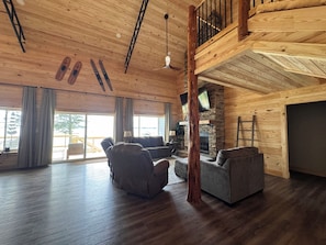 A spacious living area with a high ceiling, wooden beams, a stone fireplace, and a view through large windows. - Lake Daze . (Fort Gaines)