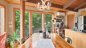 A wooden dining table with chairs, a chandelier, a kitchen area with a refrigerator, and large windows with greenery outside. - Beech Mt Place - Cool Mtn Escape & Big Views! (Beech Mountain)