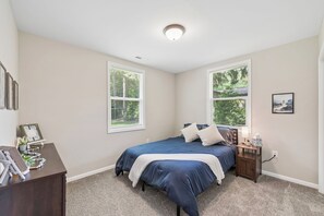 A bedroom with a bed, two windows with blinds, a bedside table with a lamp, and a small painting on the wall.