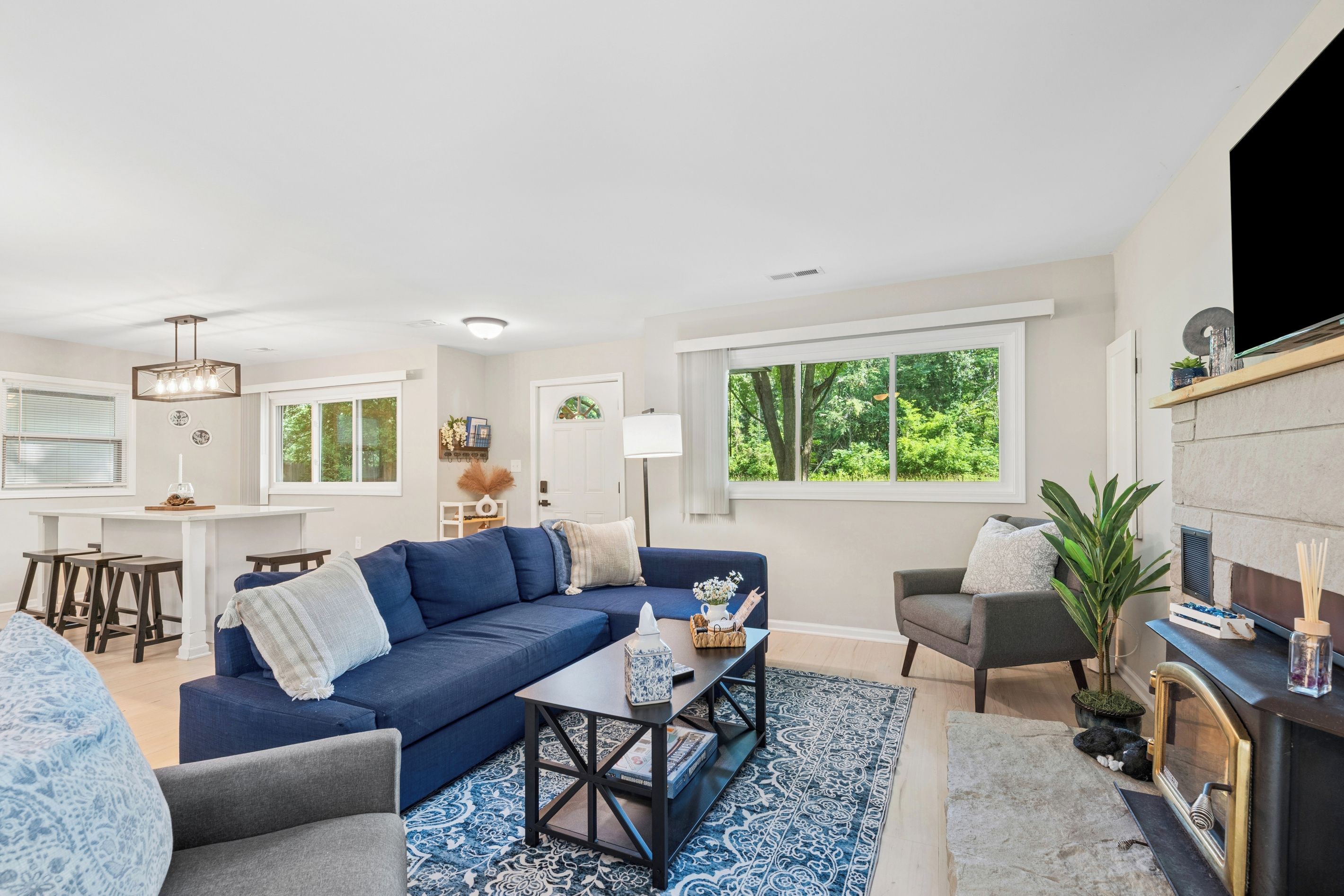 A modern living room with a blue sofa, a coffee table, and a fireplace. There is a dining area with a white table and bar stools.