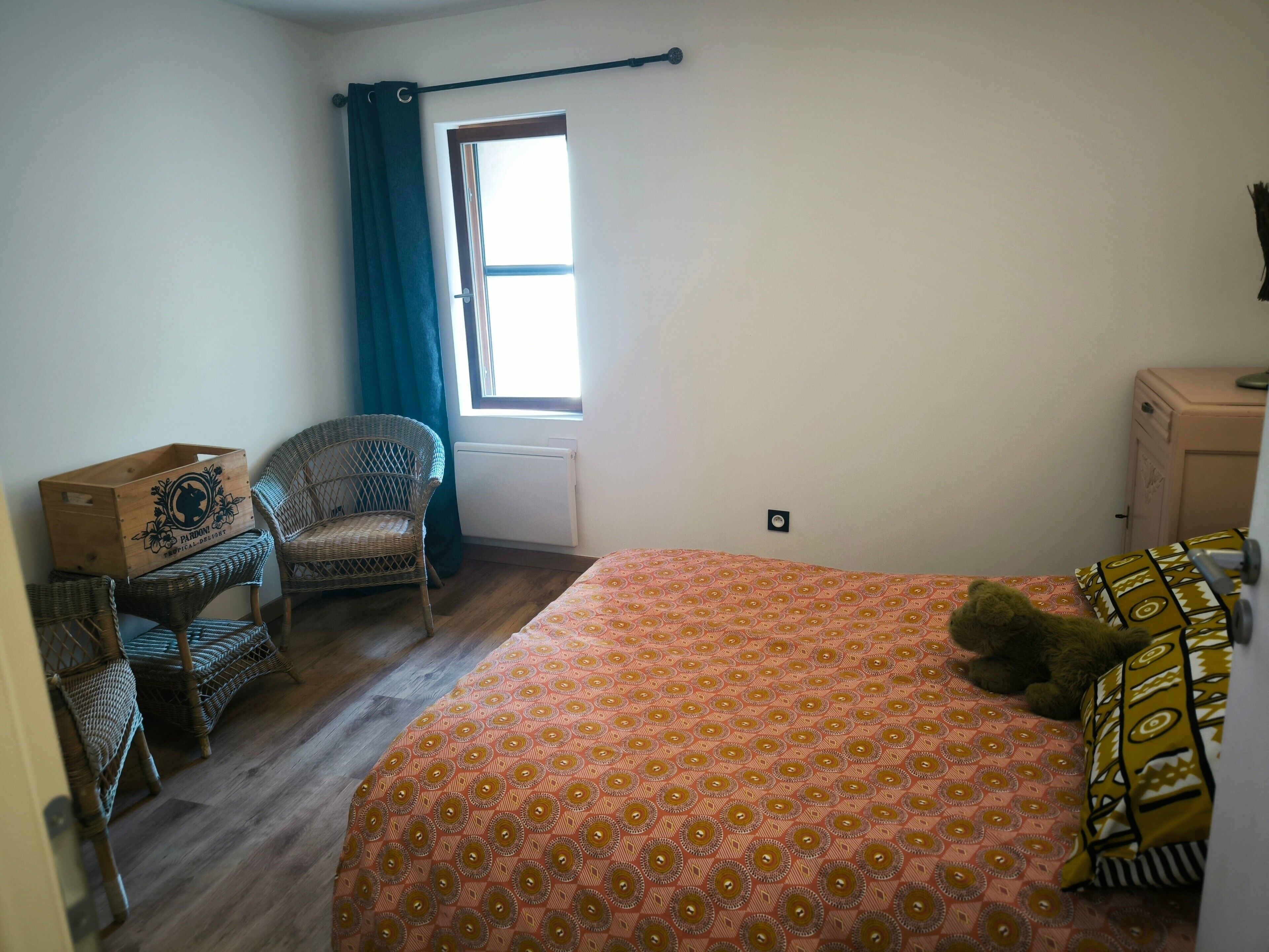 Une chambre à coucher comprenant un lit, une chaise, un radiateur, une fenêtre avec des rideaux et un petit meuble.