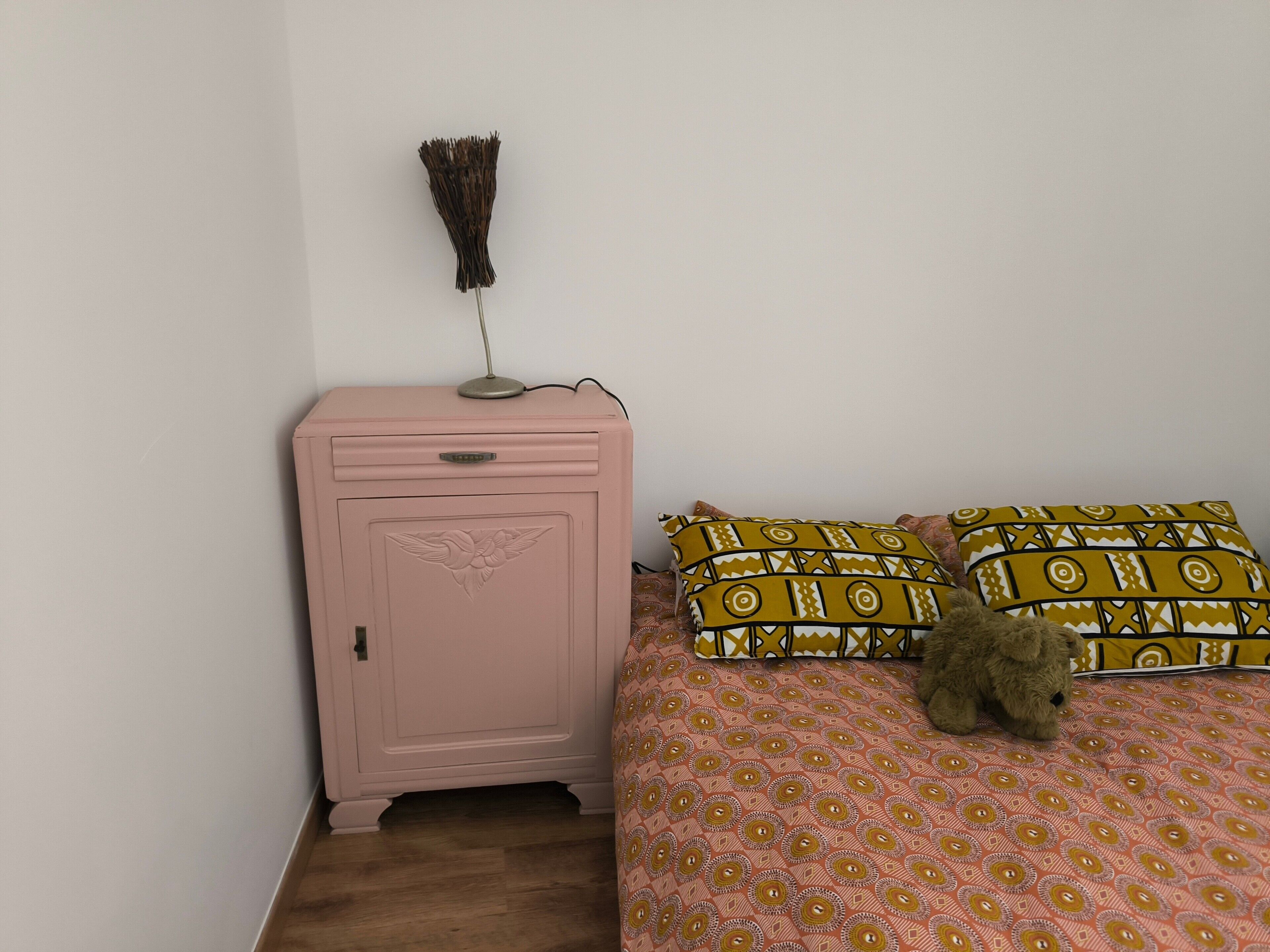 A bedroom with a pink nightstand, a lamp, a patterned bedspread, and a teddy bear.