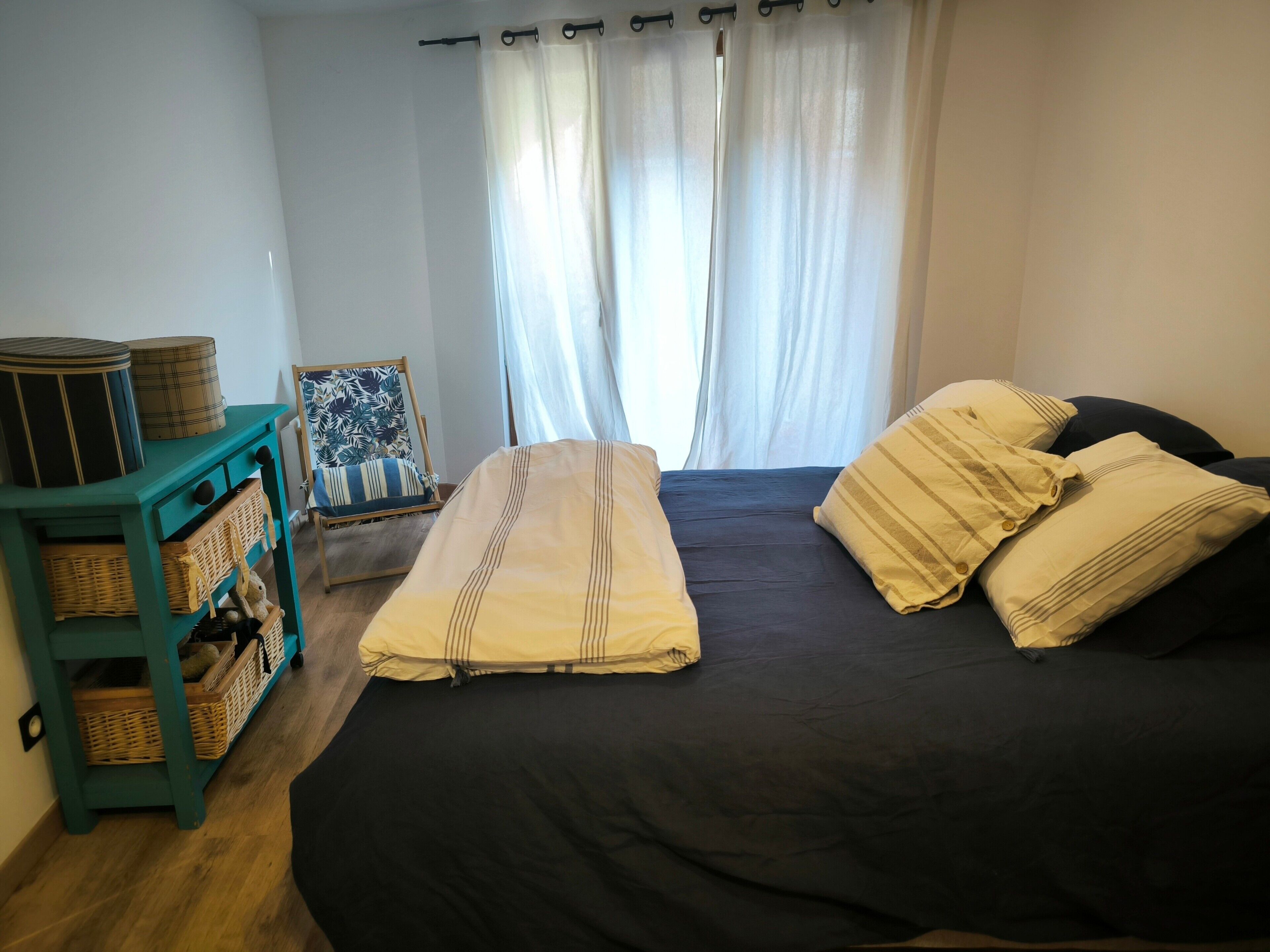 Une chambre à coucher avec un lit, des oreillers, une chaise et une petite table sur laquelle se trouvent des paniers.