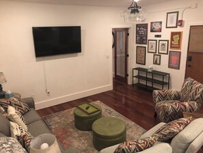 Ein Wohnzimmer mit Flachbildfernseher an der Wand, einer Couch, Hocker, einem gemusterten Teppich und einem kleinen Tisch mit einer Lampe.
