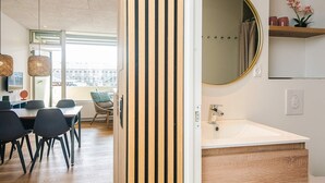 Un intérieur moderne avec un séparateur en bois, un miroir rond, un lavabo blanc et un coin repas comprenant des chaises et une table.