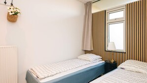 Une chambre avec deux lits simples, un radiateur, une fenêtre avec des stores verticaux et une lampe fixée au mur.