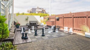 Un jeu d’échecs est installé sur une terrasse dallée, avec un banc en bois et des plantes en pot à proximité.