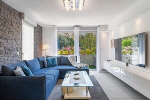 Un salon moderne avec un canapé d’angle, une table basse en verre, un grand téléviseur et une vue sur un jardin grâce à de grandes portes vitrées.