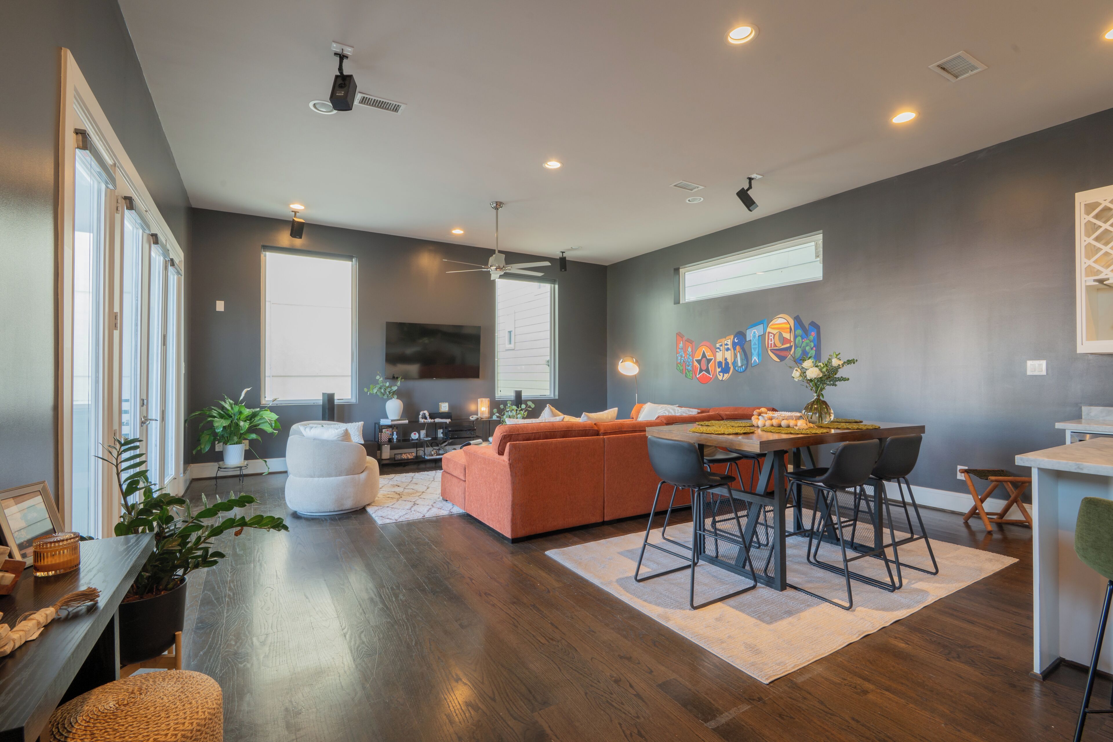 A modern living room with a sofa, dining table, and chairs, a ceiling fan, and a large window.