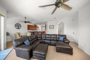 Ein geräumiges Wohnzimmer mit einer großen, L-förmigen Couch, einem Deckenventilator und einer Küchenzeile im Hintergrund.