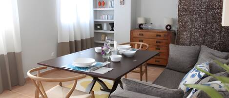 A dining area with a table set for two, a sofa, a wooden dresser, and a decorative wall piece.
