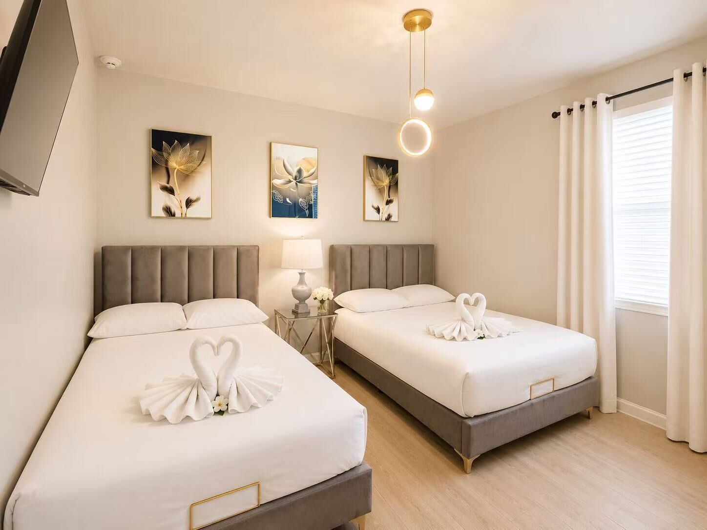 A hotel room with two beds, a TV, artwork on the wall, and a window with curtains.