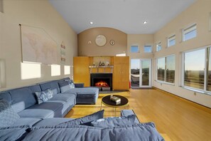 A spacious living room with a large sofa, a coffee table, and a fireplace. There are multiple windows providing natural light and a view of the outdoors. - Panoramic Estate As Seen on 90 Day Fiancé (La Mesa)