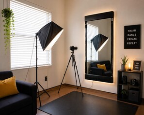 Ein Zimmer mit einer auf einem Stativ montierten Kamera, zwei Studioleuchten, einem großen Spiegel, einem schwarzen Sofa, einem kleinen Tisch mit Pflanze und einem Motivationsschild sowie einem Fenster mit Jalousien.
