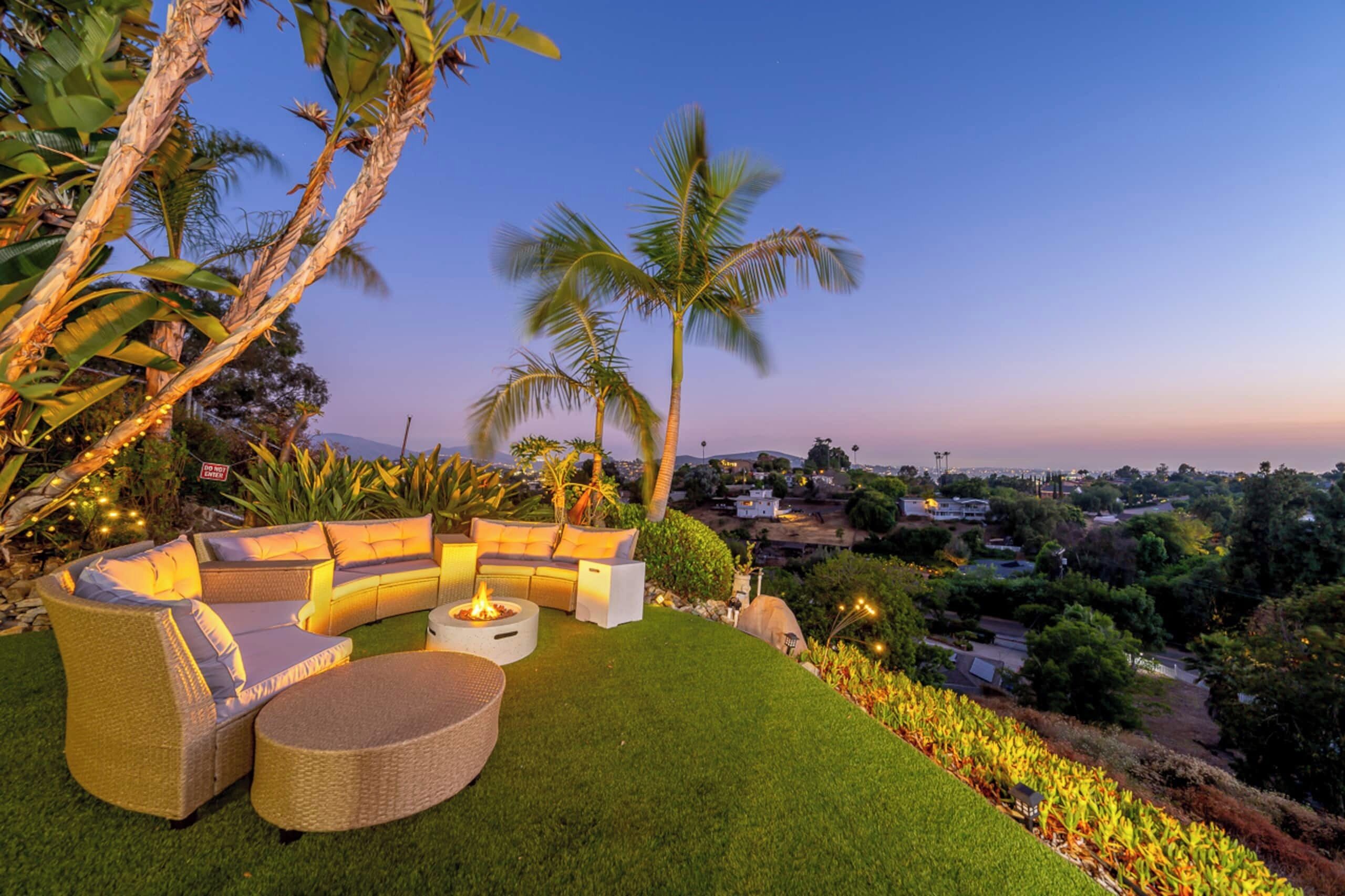 A well-maintained outdoor seating area with a curved sofa, a round table, and a fire pit, surrounded by lush greenery and palm trees.