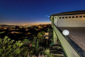 Ein Wohngebäude mit Garage, einem Balkon mit einer kugelförmigen Lampe und Blick auf eine Stadtlandschaft im Abendlicht.