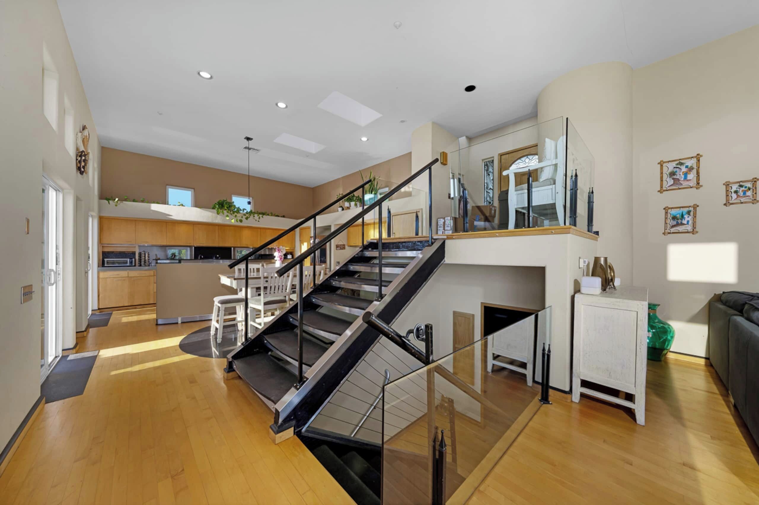 A spacious living area with a modern staircase, a kitchen in the background, and a dining area with a glass railing.