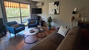 A living room with a sofa, two armchairs, a coffee table, a TV, a plant, and a large window. - Canal & Garonne Atmosphere – Live Like a True Toulousain (Toulouse)