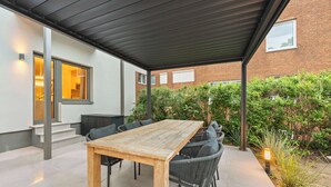 Eine überdachte Terrasse mit einem Holztisch und schwarzen Korbsesseln, ein Backsteinhaus im Hintergrund und ein kleiner Gartenbereich mit einer Lampe.