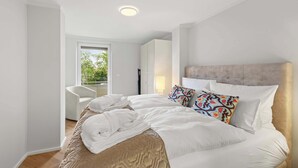 Ein modernes Hotelzimmer mit einem großen Bett, einem Sessel, einem Kleiderschrank und Blick auf Grünfläche.
