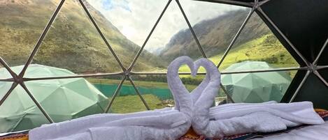Una habitación con un diseño de ventana geométrico, una cama con una toalla en forma de corazón y vistas a un paisaje montañoso.