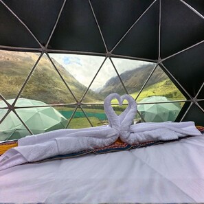 Ein Zimmer mit einem geometrisch gestalteten Fenster, einem Bett mit einem herzförmigen Handtuch und Blick auf eine gebirgige Landschaft.