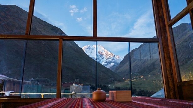 Una habitación con un ventanal grande que ofrece vistas a la montaña, una mesa de madera y un tapete estampado.