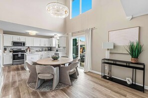 A modern kitchen with white cabinetry, a dining area with a round table and chairs, and a large window with curtains.