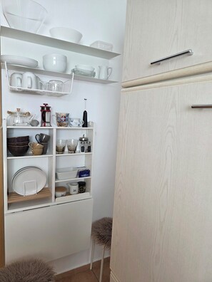 A kitchen with white cabinetry, shelves with dishes, a coffee maker, and a stool. - Vacation rental near Sylt with a balcony and parking in the heart of Leck (Leck)
