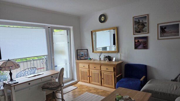 Una habitación con escritorio, silla y mesa de comedor con una bandeja de objetos. Hay una silla azul, un armario de madera y un espejo en la pared.