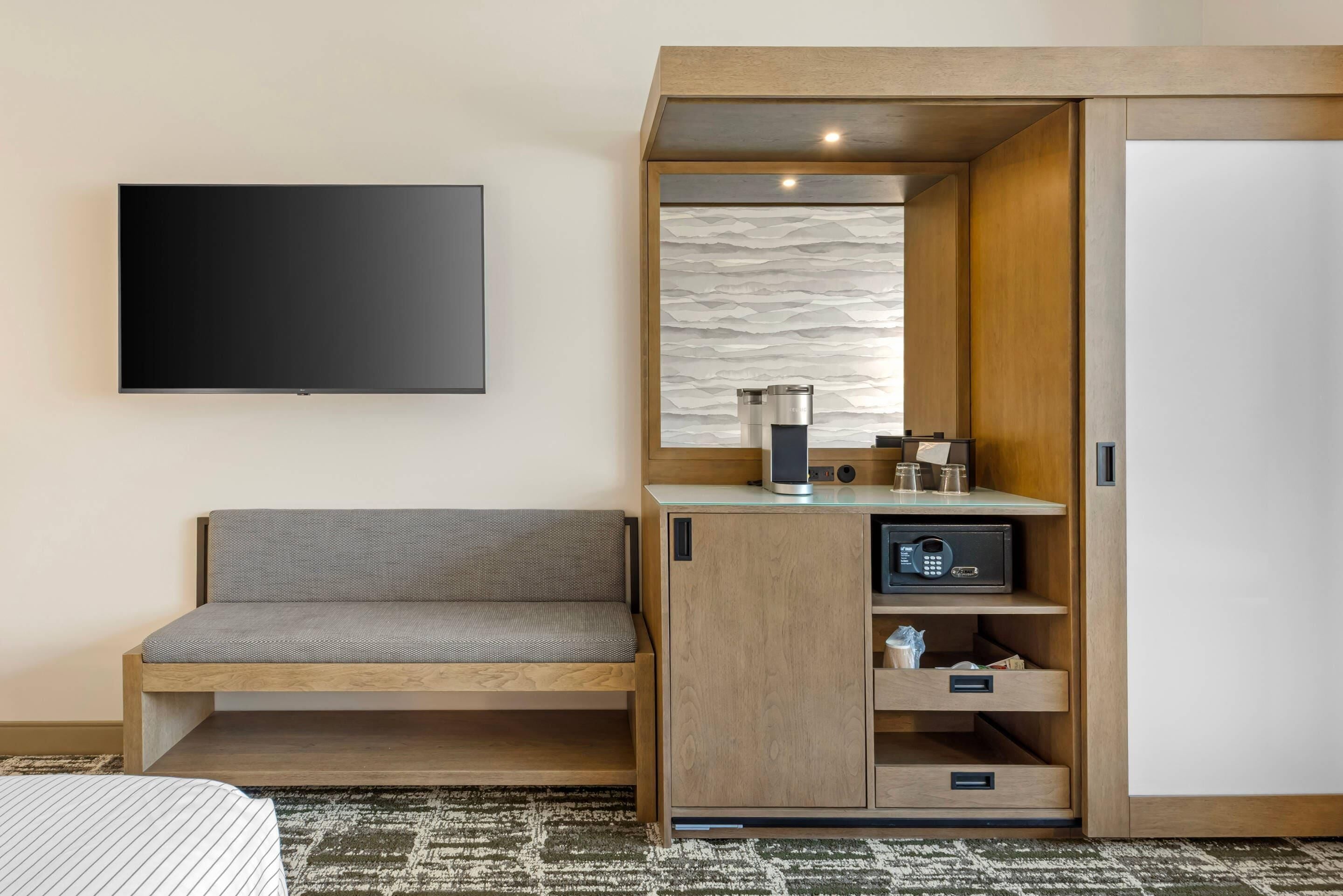 A hotel room with a flat-screen TV mounted on the wall, a sofa, a wooden cabinet with a coffee maker, and a minibar.