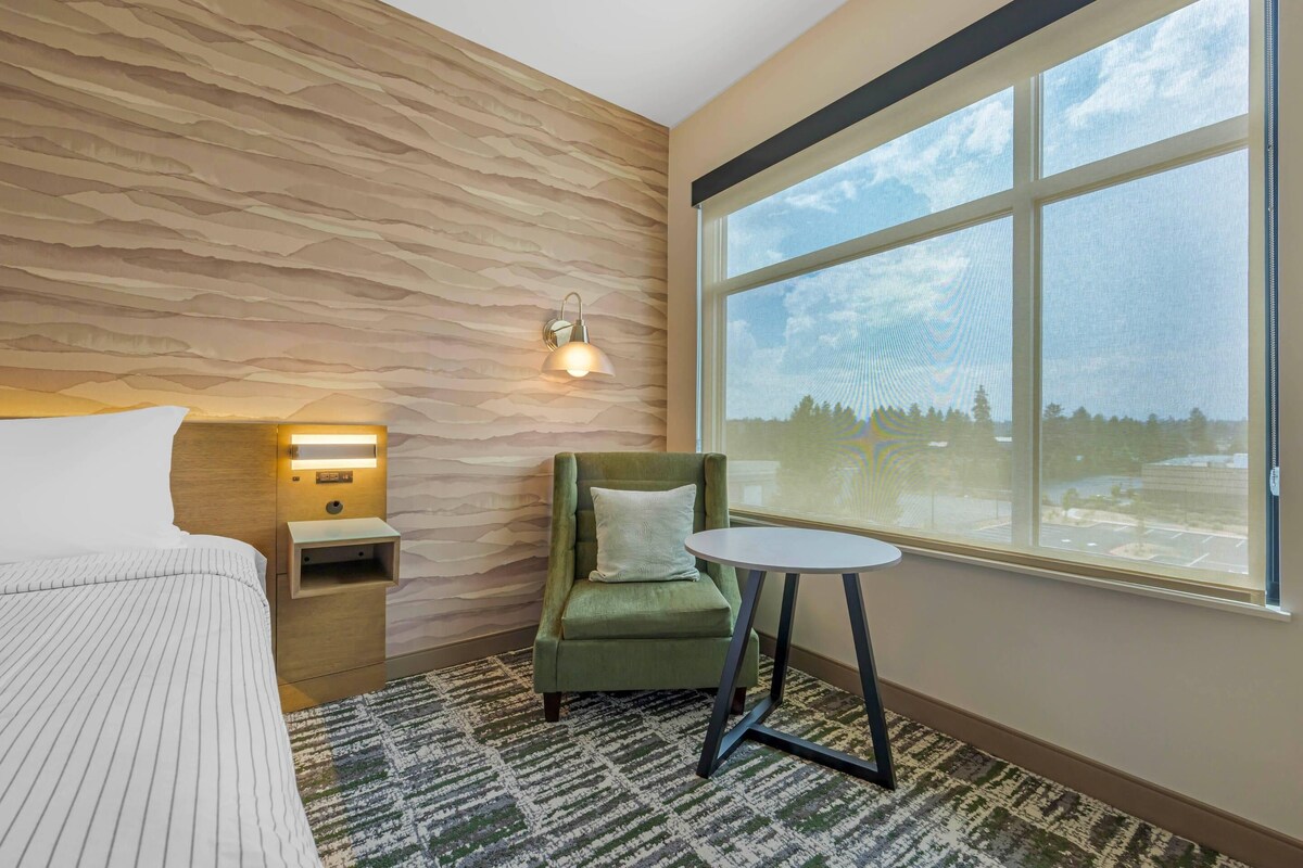 A hotel room with a bed, a green armchair, a small table, a nightstand, a wall-mounted lamp, and a large window with a view.