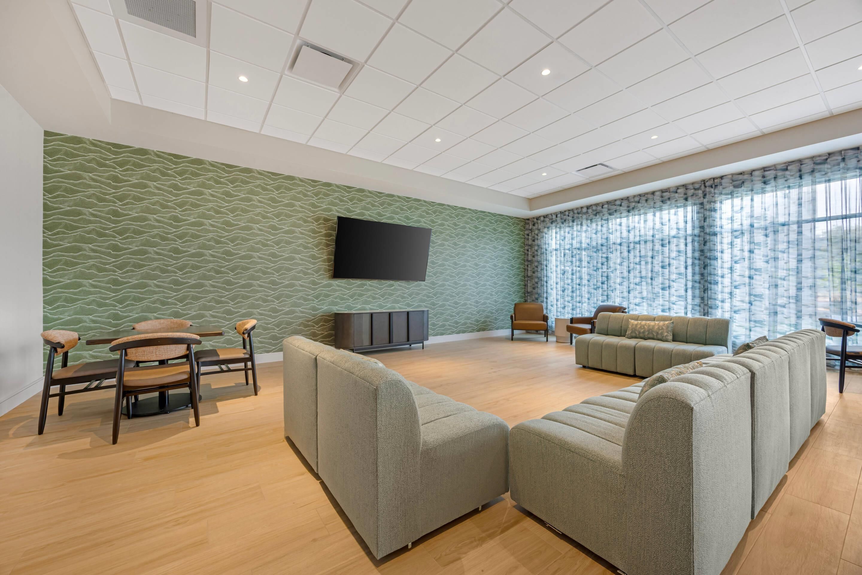 A spacious living area with a green textured wall, a flat-screen TV mounted above a dark-colored entertainment unit, and a large window with sheer curtains.