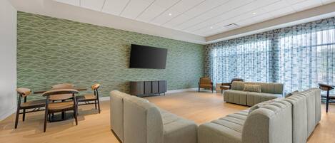 A spacious living area with a green textured wall, a flat-screen TV mounted above a dark-colored entertainment unit, and a large window with sheer curtains.