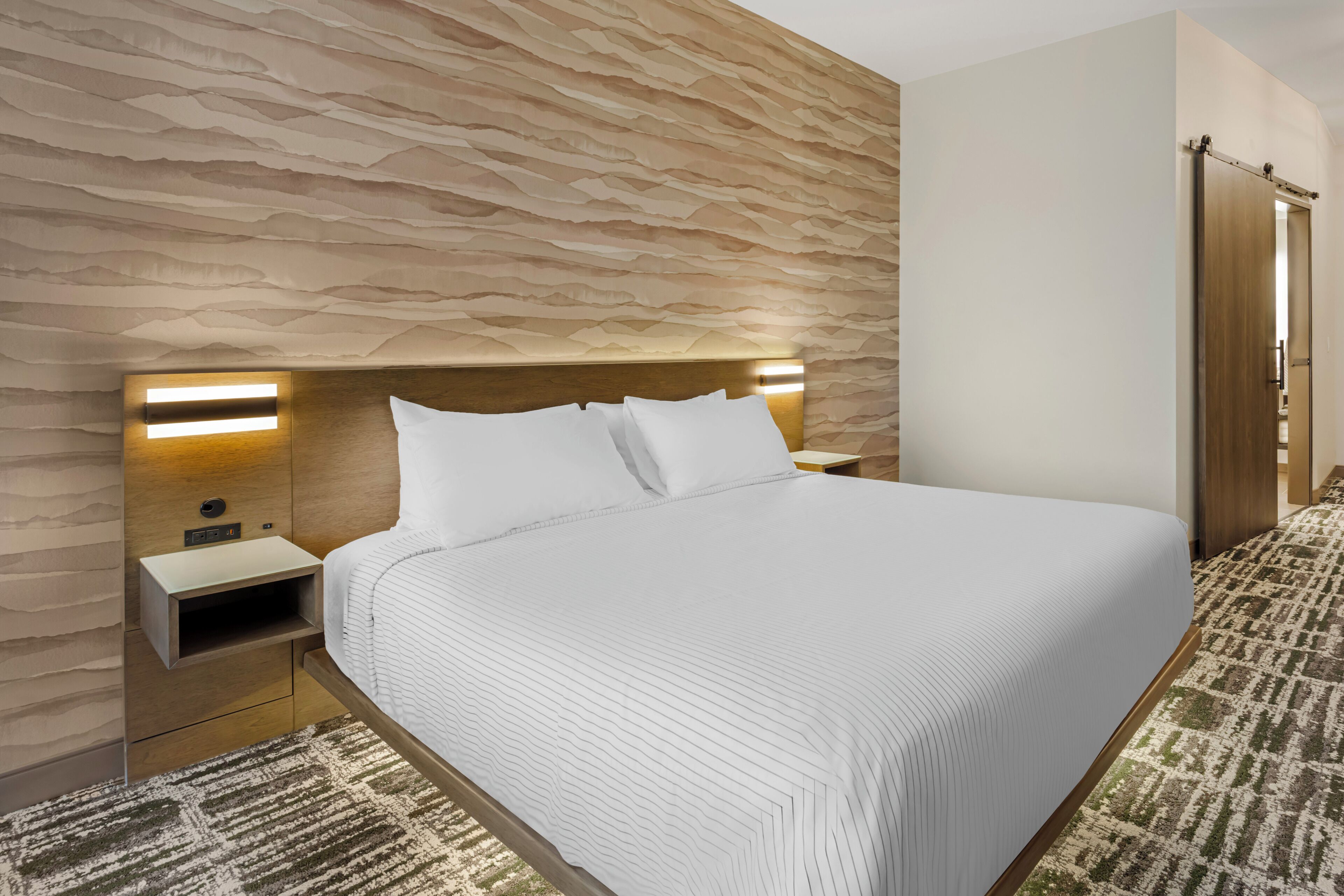 A modern hotel room with a large bed, a wooden headboard, bedside lamps, a nightstand, and a patterned carpet.