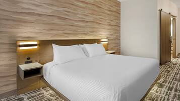 A modern hotel room with a large bed, a wooden headboard, bedside lamps, a nightstand, and a patterned carpet.