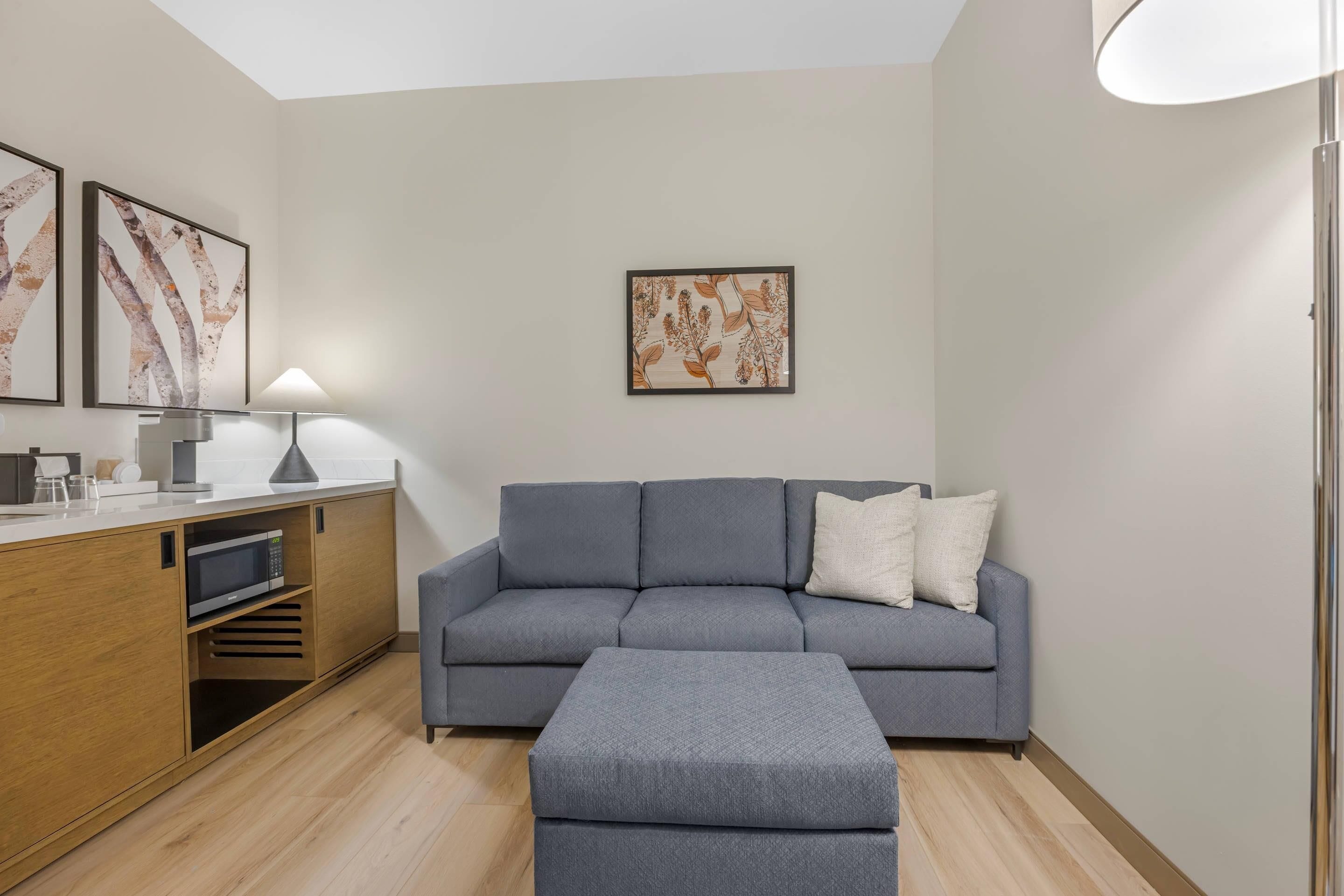 Una sala moderna con un sofá gris, un pequeño reposapiés, un mueble de madera con microondas y dos lámparas de pared.