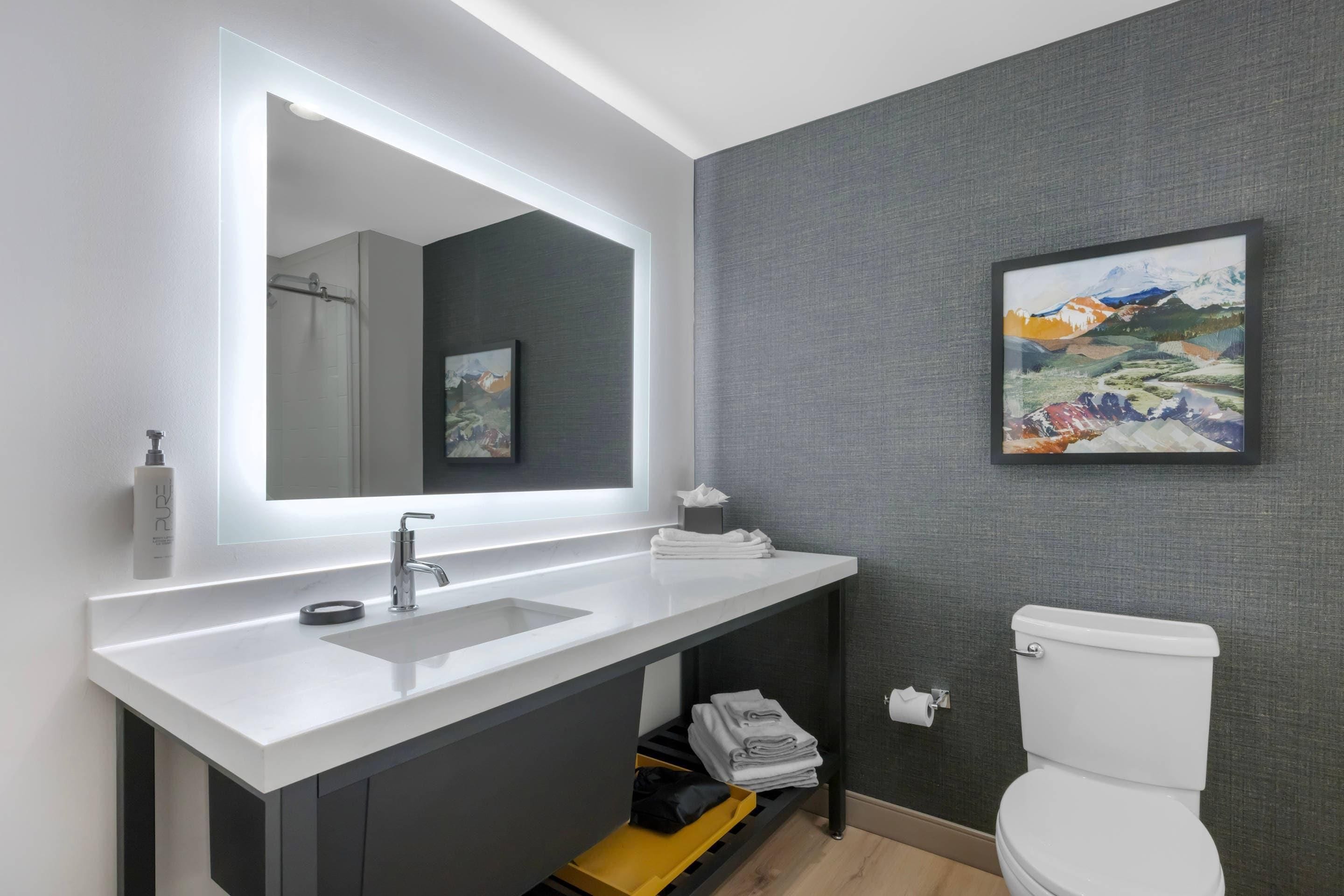 Uma casa de banho moderna com uma pia branca, um espelho grande, uma sanita e um quadro decorativo com uma paisagem.
