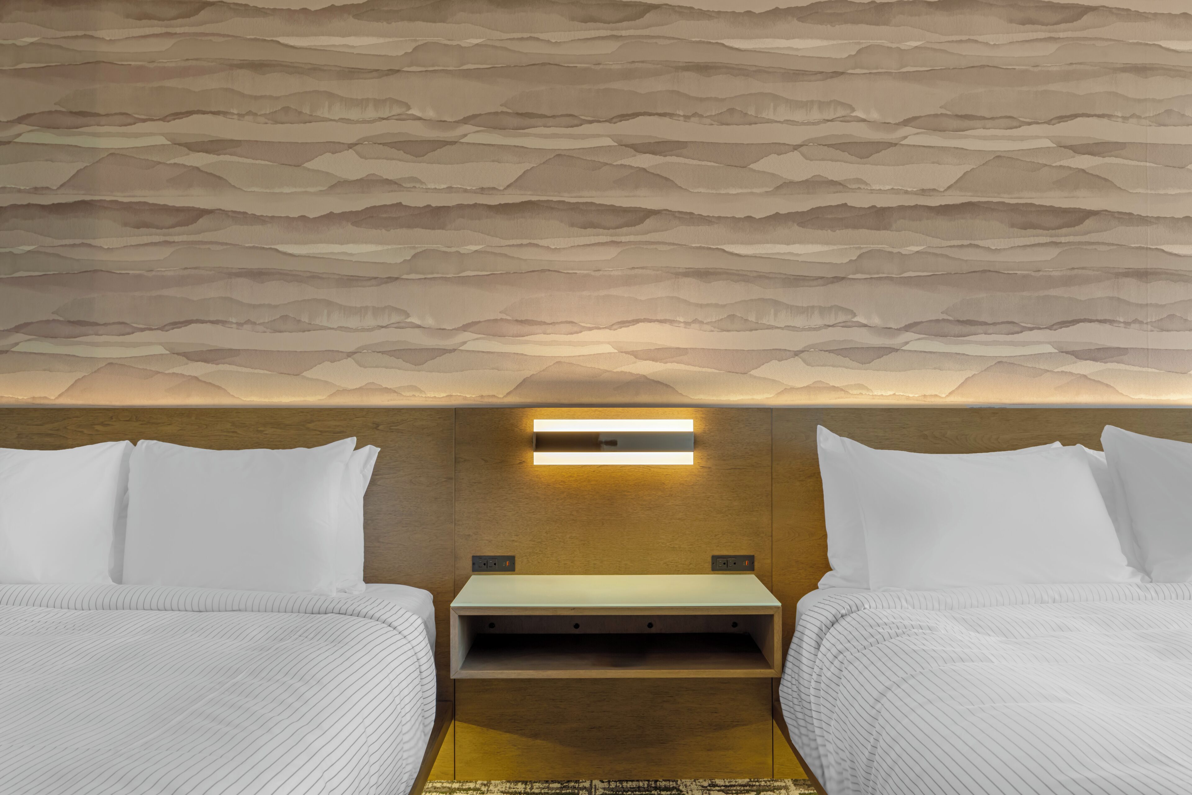 Habitación de hotel con techo texturizado, una mesita de noche, dos camas con ropa blanca y una lámpara fijada a la pared.