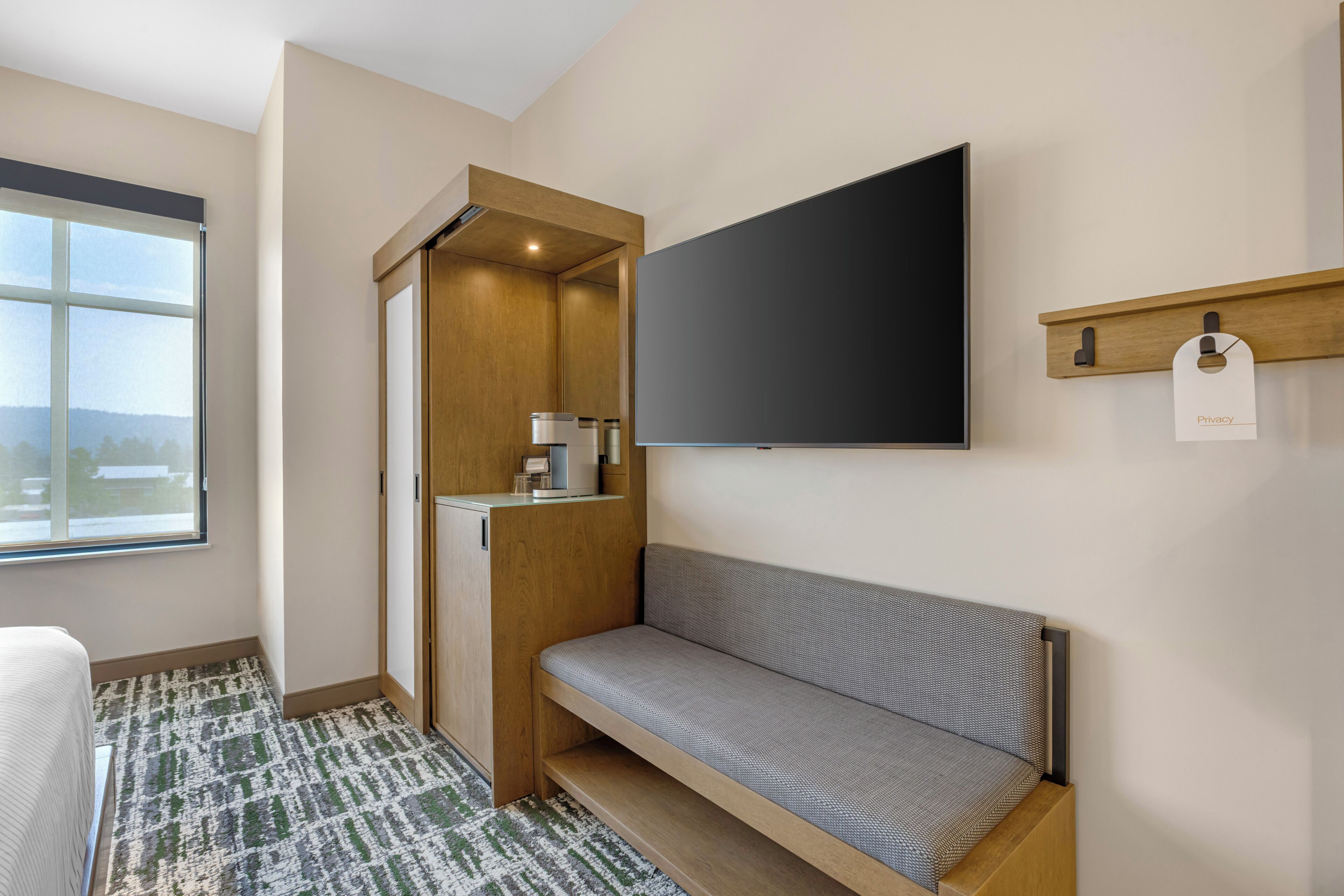 A modern hotel room with a large window, a built-in bench, a flat-screen TV mounted on the wall, and a small shelf with a privacy tag.