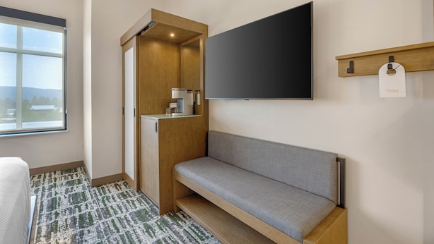 A modern hotel room with a large window, a built-in bench, a flat-screen TV mounted on the wall, and a small shelf with a privacy tag.
